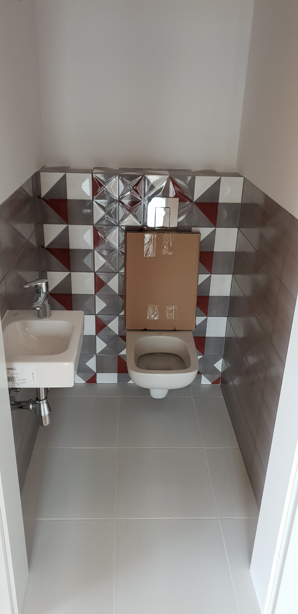 Mała łazienka z umywalką i toaletą, ściany wyłożone płytkami w geometryczne wzory, toaleta zakryta kartonem ochronnym.