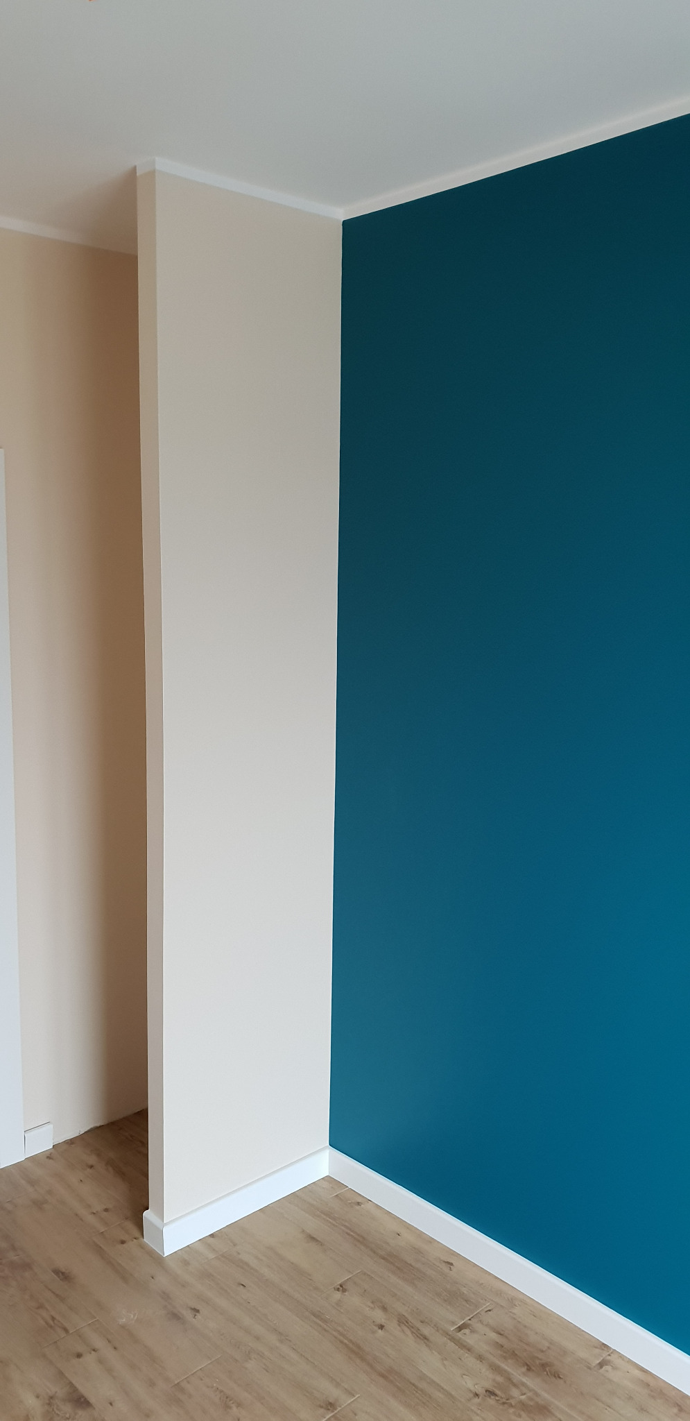 Pomieszczenie po malowaniu: ściana w odcieniu petrolu kontrastuje z beżową ścianą i drewnopodobną podłogą, widoczne białe listwy przypodłogowe.