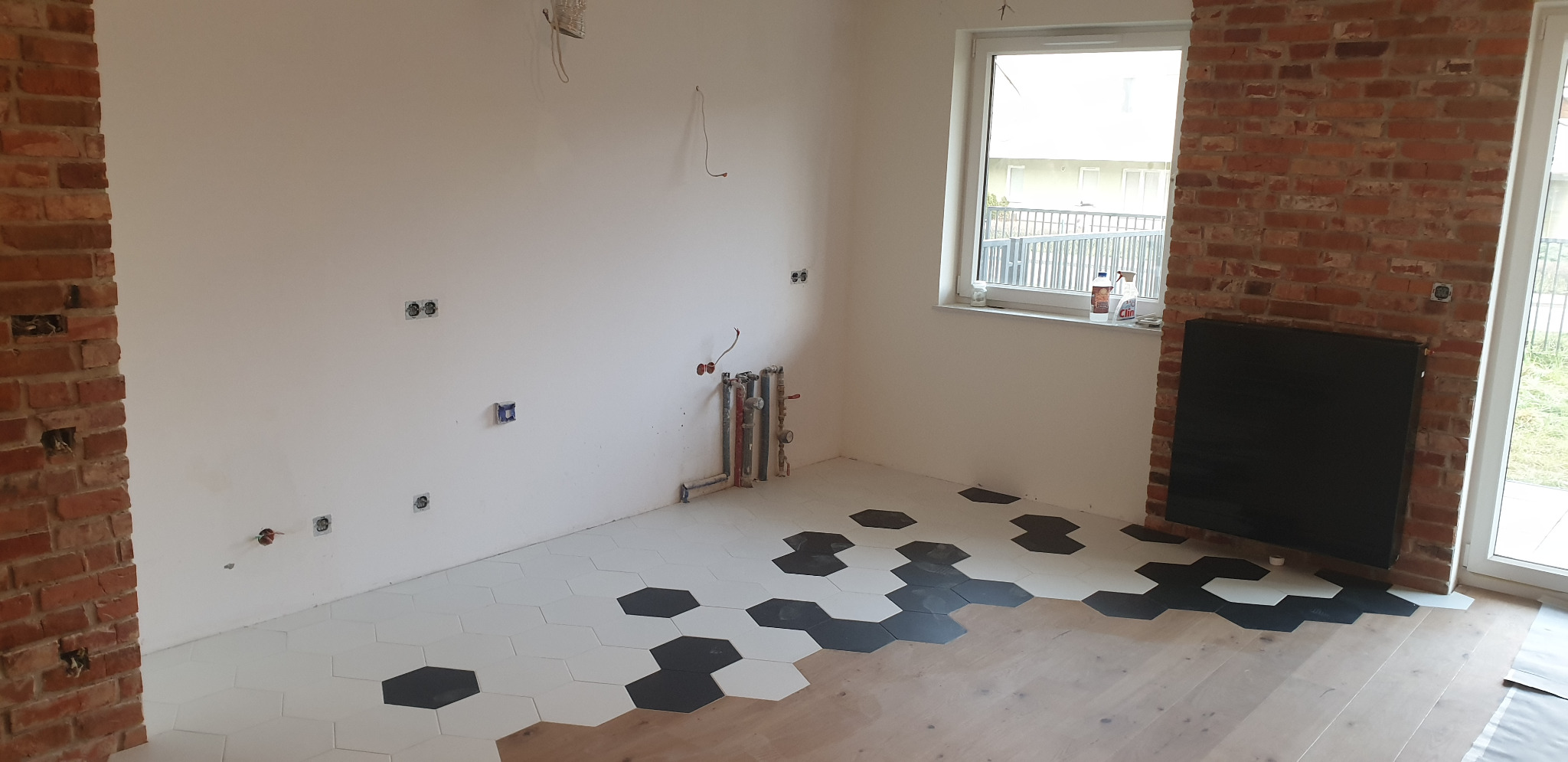 Wnętrze w trakcie remontu: kontrastowe heksagonalne płytki podłogowe (białe i czarne) przechodzące w drewniany parkiet, ściany z instalacją elektryczną, fragment ściany z cegły, okno z widokiem...