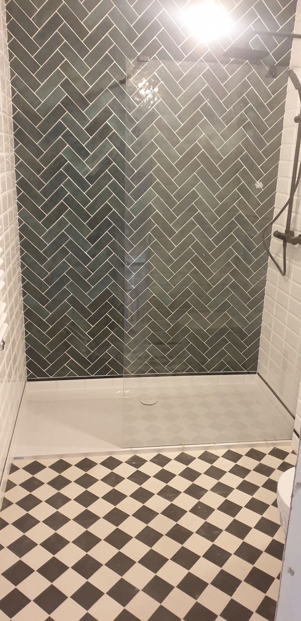 Nowoczesna kabina prysznicowa z ciemnozielonymi płytkami ułożonymi w jodełkę na ścianie, szklaną ścianką i podłogą w czarno-białą szachownicę.