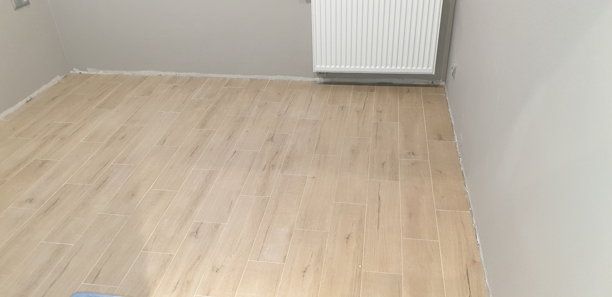 Ułożona podłoga z płytek imitujących drewno w jasnym odcieniu, widoczny grzejnik na ścianie i szara ściana, świeżo po fugowaniu.
