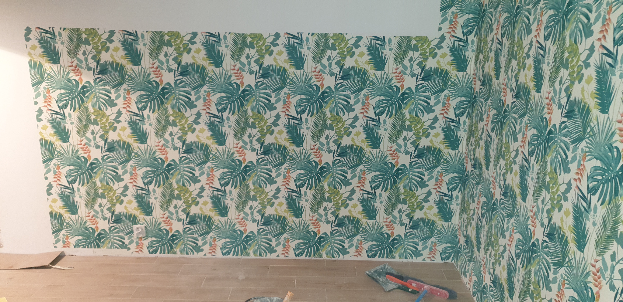 Ściana i róg pokoju pokryte tapetą w tropikalne liście, widoczne narzędzia i fragment podłogi w trakcie prac wykończeniowych.
