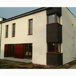 Nowoczesny dom z białą elewacją i ciemnymi elementami dekoracyjnymi okiennymi. Widoczne brązowe ramy okien i częściowo wykończony teren wokół budynku.