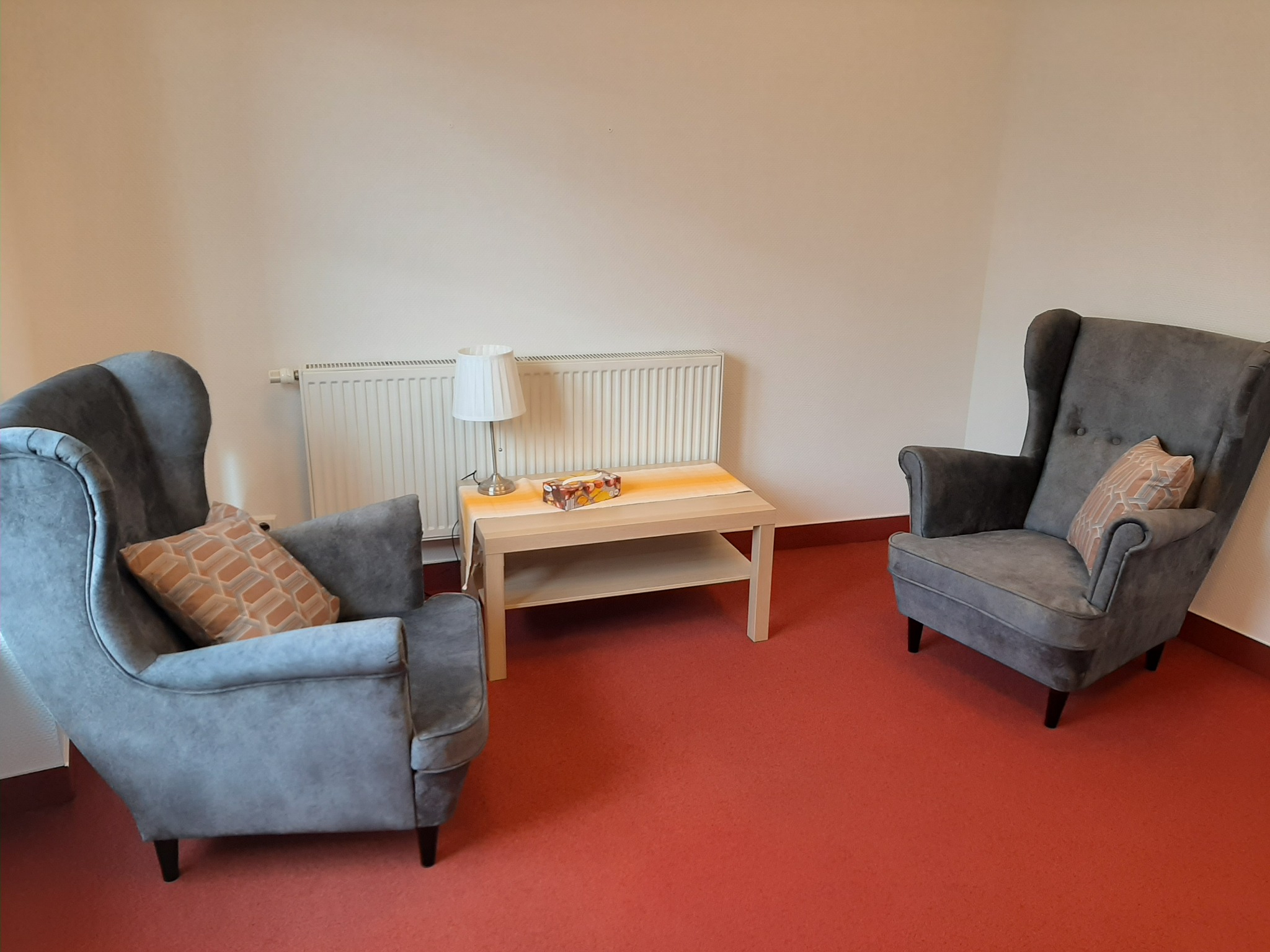 Pokój z dwoma szarymi fotelami uszakami z poduszkami w geometryczne wzory, stolikiem kawowym z lampką i pudełkiem chusteczek, na czerwonej wykładzinie.