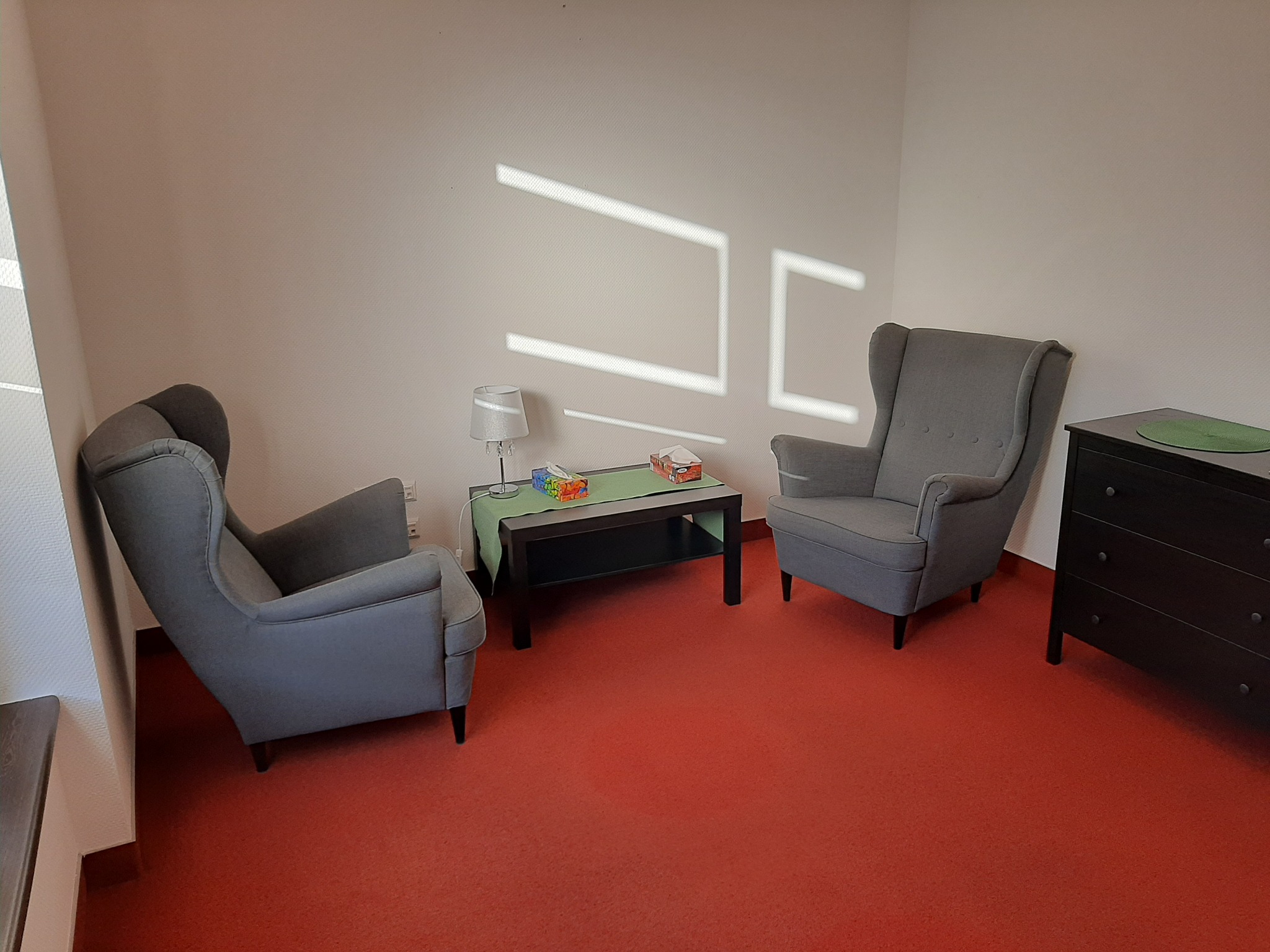 Pokój z dwoma szarymi fotelami uszakami, małym stolikiem z lampką i pudełkiem chusteczek, oraz komodą z zieloną podkładką, oświetlony promieniami słońca wpadającymi przez okno.