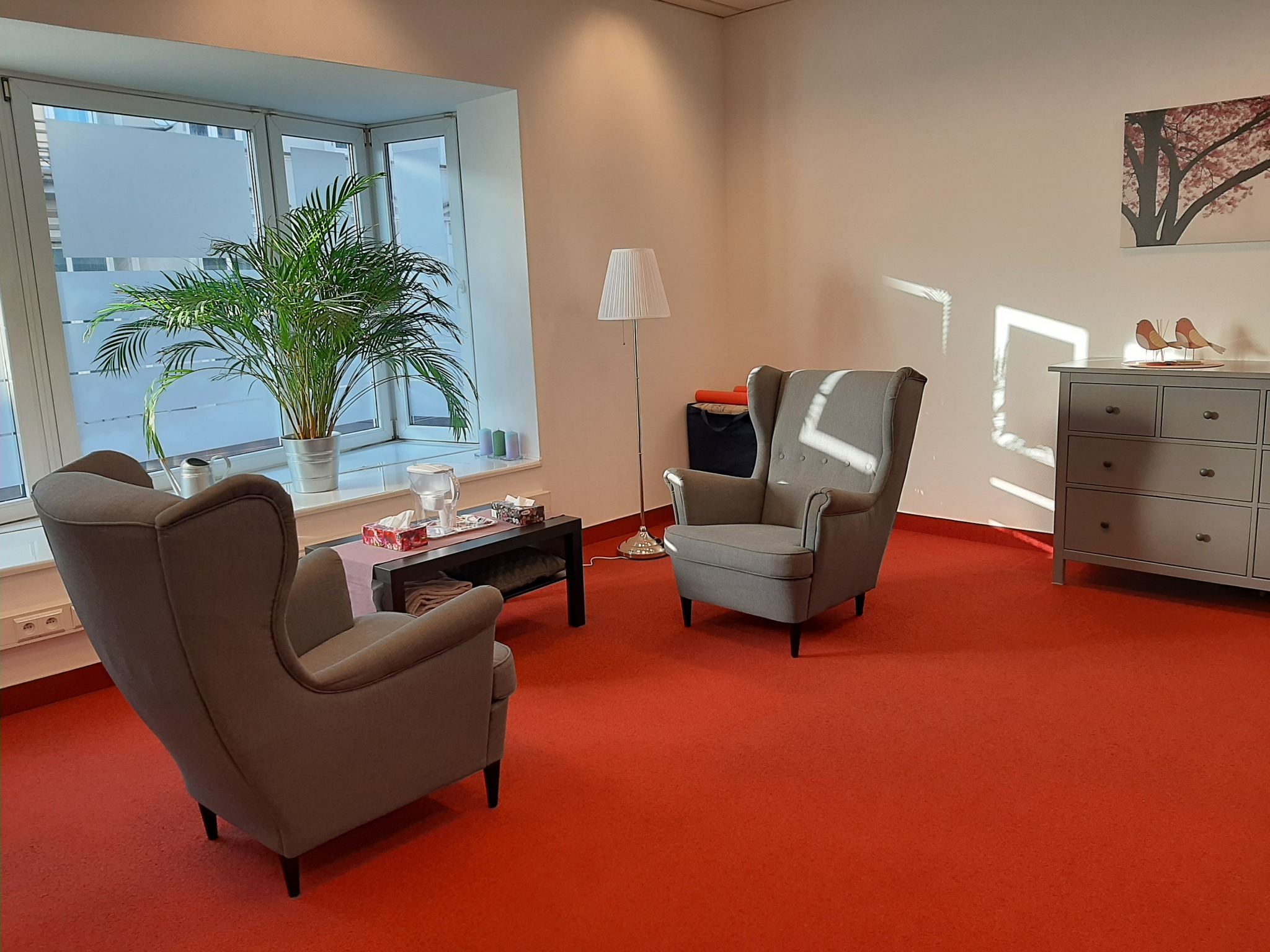Gabinet terapeutyczny z dwoma szarymi fotelami typu uszak, stolikiem kawowym z chusteczkami, lampą podłogową i komodą z dekoracją w postaci ptaków, oświetlony światłem dziennym wpadającym przez...