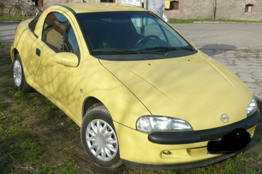 Żółty Opel Tigra z 1997 roku, sfotografowany na zewnątrz w słoneczny dzień, z widocznym rowerem w tle i elementami muru.