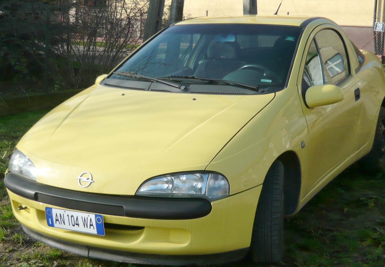 Żółty Opel Tigra z 1997 roku, zaparkowany na trawie przed budynkiem z numerem 20, widoczny numer rejestracyjny AN 104 WA.