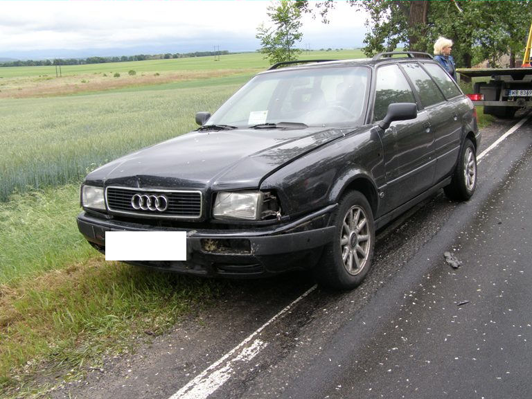 Czarny uszkodzony Audi 80 Avant z 1992 roku po kolizji, stojący na poboczu drogi z widocznymi uszkodzeniami przodu i brakiem tablicy rejestracyjnej, laweta w tle.