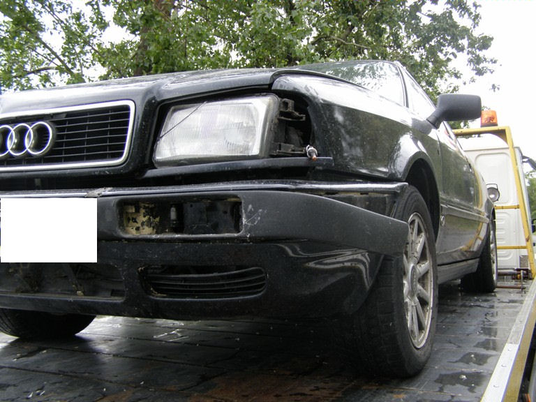 Uszkodzony przód czarnego Audi 80 Avant z 1992 roku, widoczne uszkodzenia reflektora i zderzaka, samochód na lawecie pomocy drogowej.