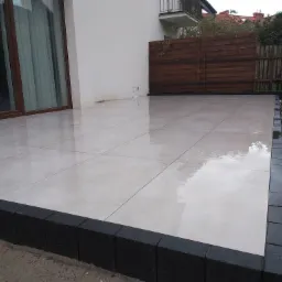 Jasne, kwadratowe płyty tarasowe z ciemnoszarą obwódką, przylegające do ściany domu z oknem tarasowym, widoczne fragmenty ogrodu i drewnianego płotu w tle.