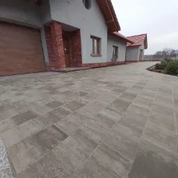 Szeroka, wybrukowana kostką ścieżka prowadząca do domu z garażem, z obrzeżem z drobnego żwiru, w tle widoczny częściowo ogród i pochmurne niebo.