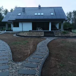Dwie ścieżki z płyt betonowych i otoczaków prowadzące do nowoczesnego domu z panelami słonecznymi na dachu, widok z perspektywy ogrodu.