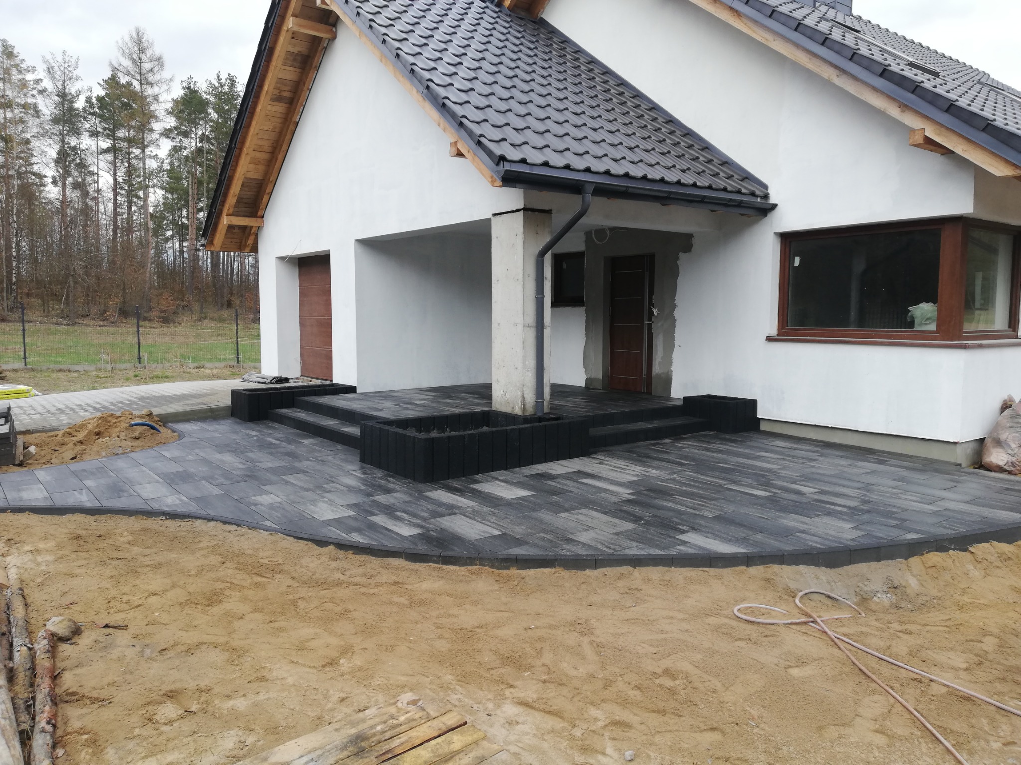 Nowo ułożony szary chodnik z kostki brukowej prowadzący do wejścia domu z ciemnym dachem, widoczne niedokończone prace ziemne.