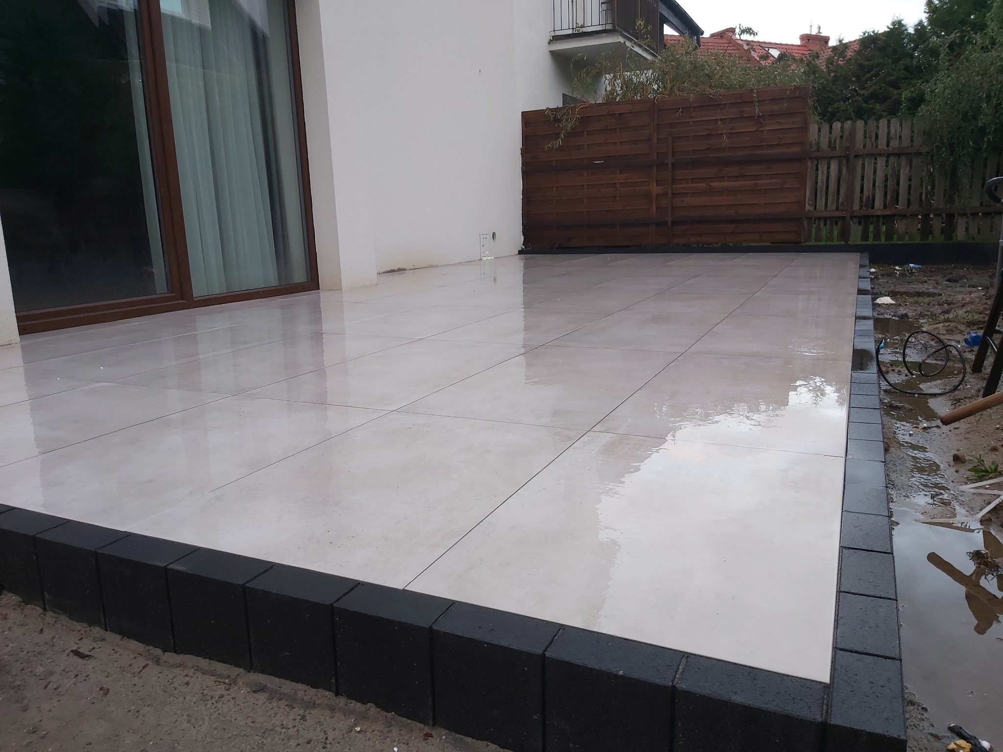 Jasne, kwadratowe płyty tarasowe z ciemnoszarą obwódką, przylegające do ściany domu z oknem tarasowym, widoczne fragmenty ogrodu i drewnianego płotu w tle.