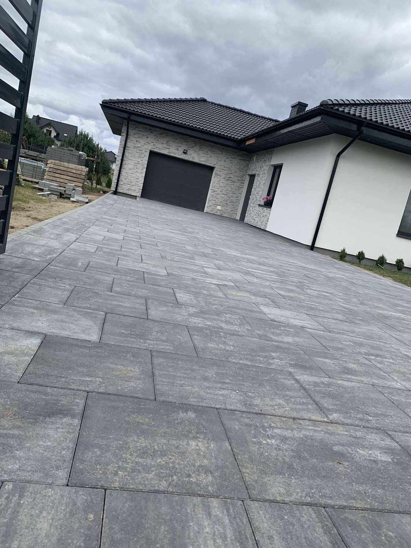 Nowoczesny podjazd z szarej kostki brukowej prowadzący do domu z garażem, widoczne ogrodzenie i przygotowane materiały budowlane.