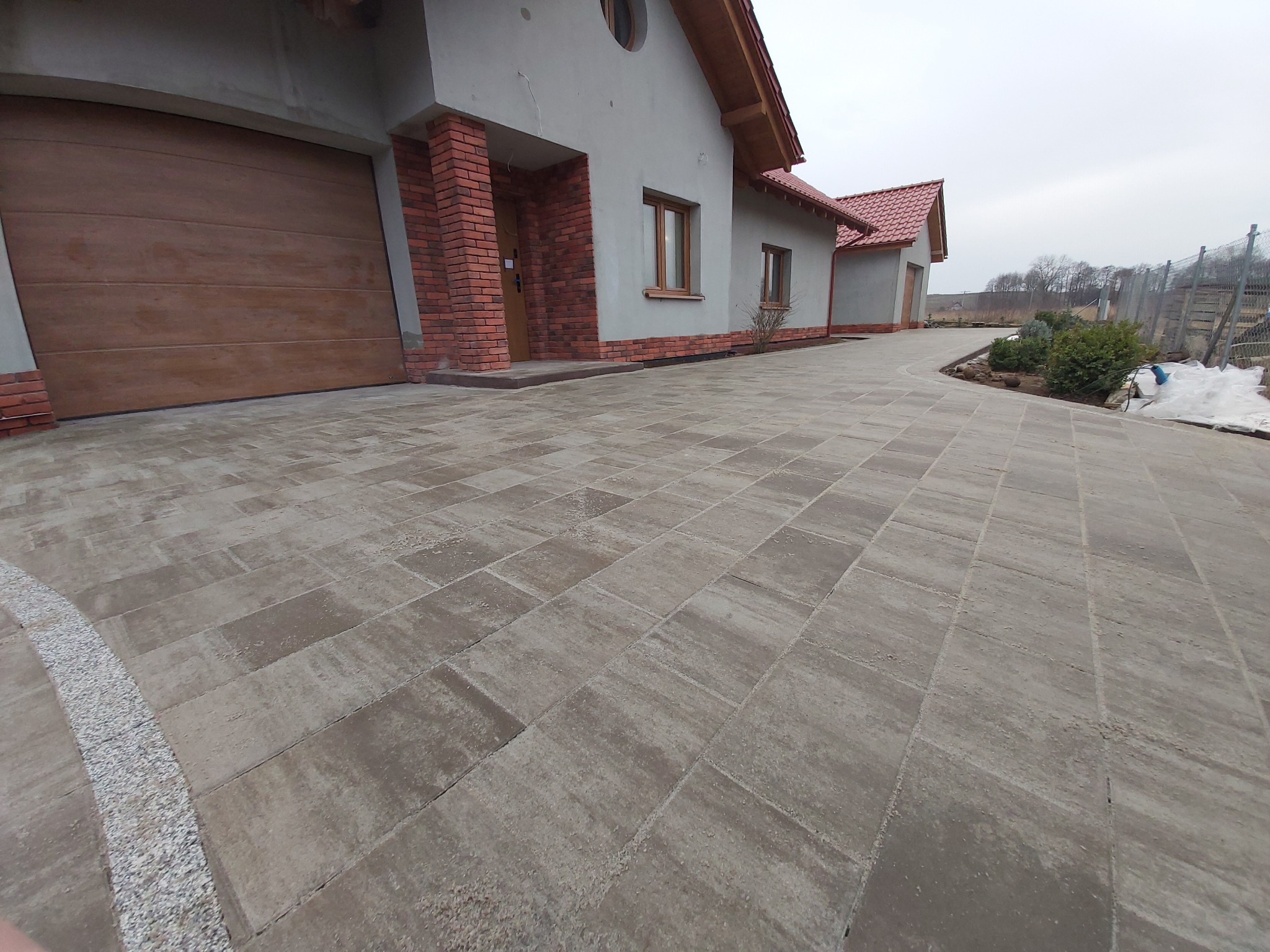 Szeroka, wybrukowana kostką ścieżka prowadząca do domu z garażem, z obrzeżem z drobnego żwiru, w tle widoczny częściowo ogród i pochmurne niebo.