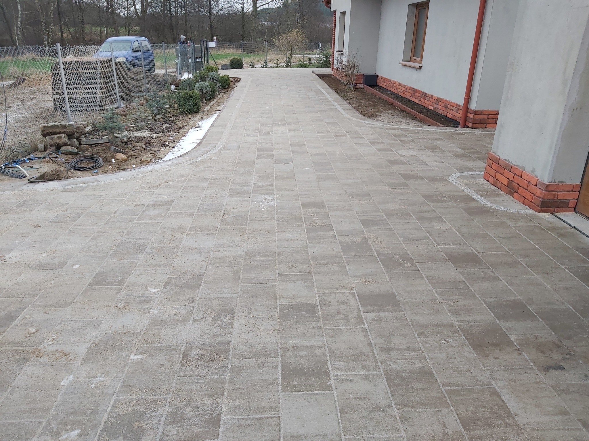 Szeroki, wybrukowany podjazd z szarej kostki prowadzący do domu z ceglaną podmurówką, widoczne świeżo posadzone krzewy i trawnik wzdłuż krawędzi.