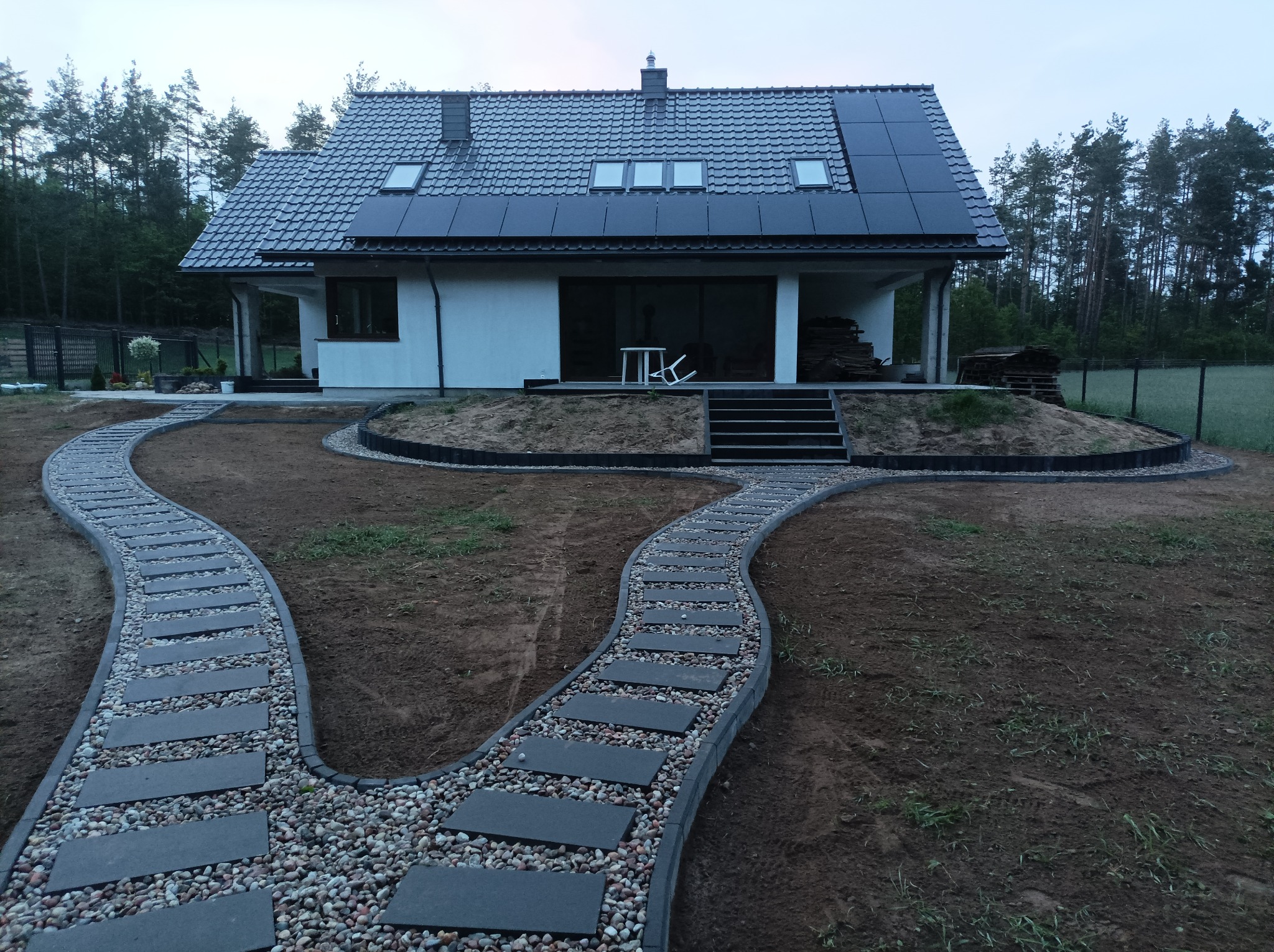 Dwie ścieżki z płyt betonowych i otoczaków prowadzące do nowoczesnego domu z panelami słonecznymi na dachu, widok z perspektywy ogrodu.