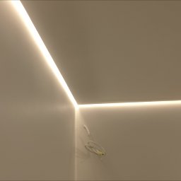 B Home Marcin Błasiak - Nowoczesne oświetlenie LED w rogu pomieszczenia, listwy świetlne tworzą geometryczny wzór na styku ściany i sufitu. Widoczne przewody elektryczne.