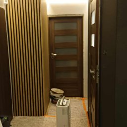 B Home Marcin Błasiak - Korytarz po remoncie w Warszawie: drewniane lamele na ścianie, podświetlany sufit, nowe drzwi. Widoczne zabezpieczenia podłogi i sprzęt budowlany.