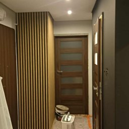 B Home Marcin Błasiak - Korytarz po remoncie: drewniane lamele na ścianie, drzwi z przeszkleniami, pomalowana ściana na ciemny kolor. Widoczny pochłaniacz wilgoci i wiaderko po farbie.