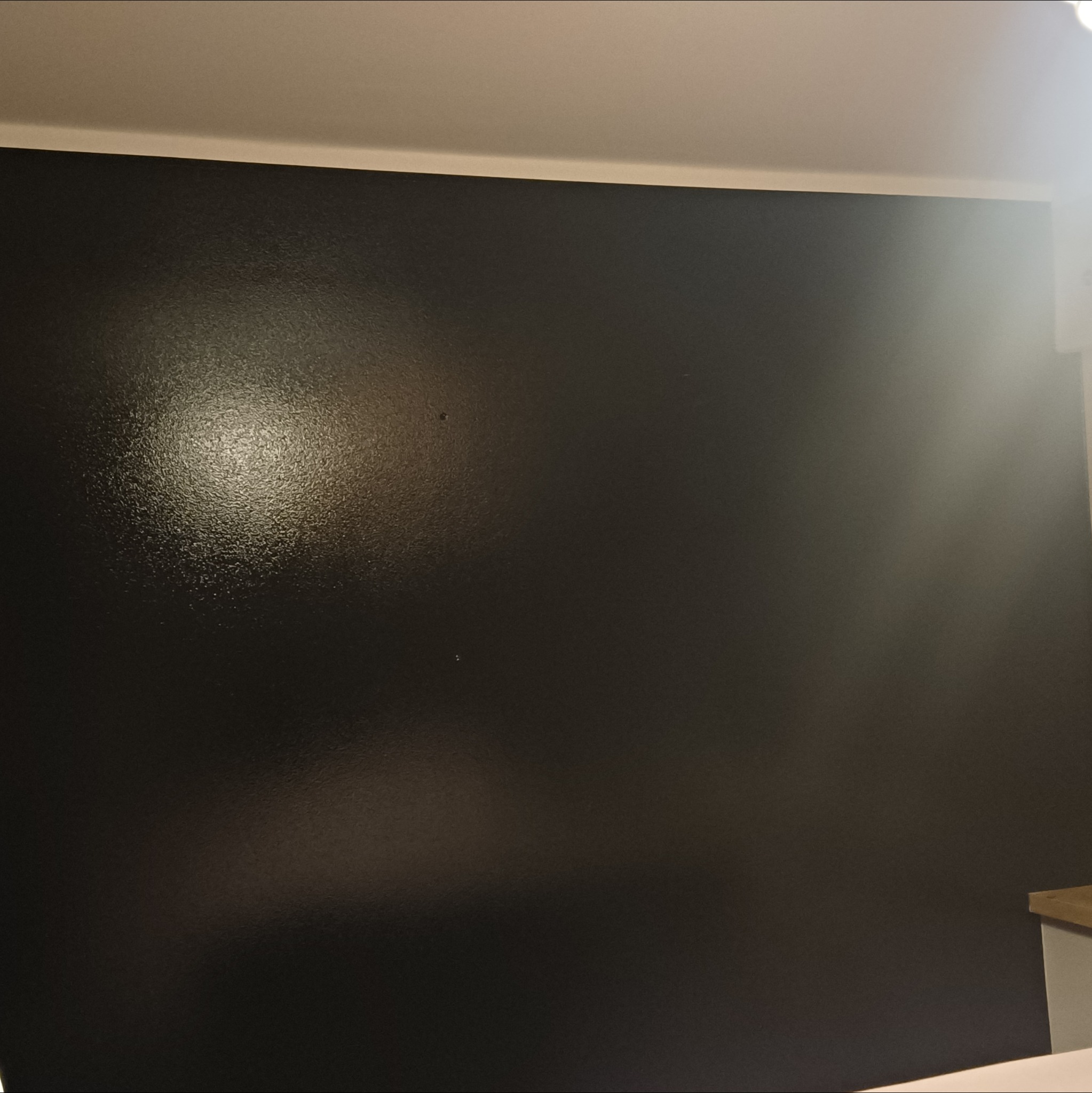 Ściana pomalowana na czarno z widoczną fakturą farby, oświetlona z góry, z fragmentem sufitu i blatu. Minimalistyczne ujęcie detalu wykończenia wnętrza.