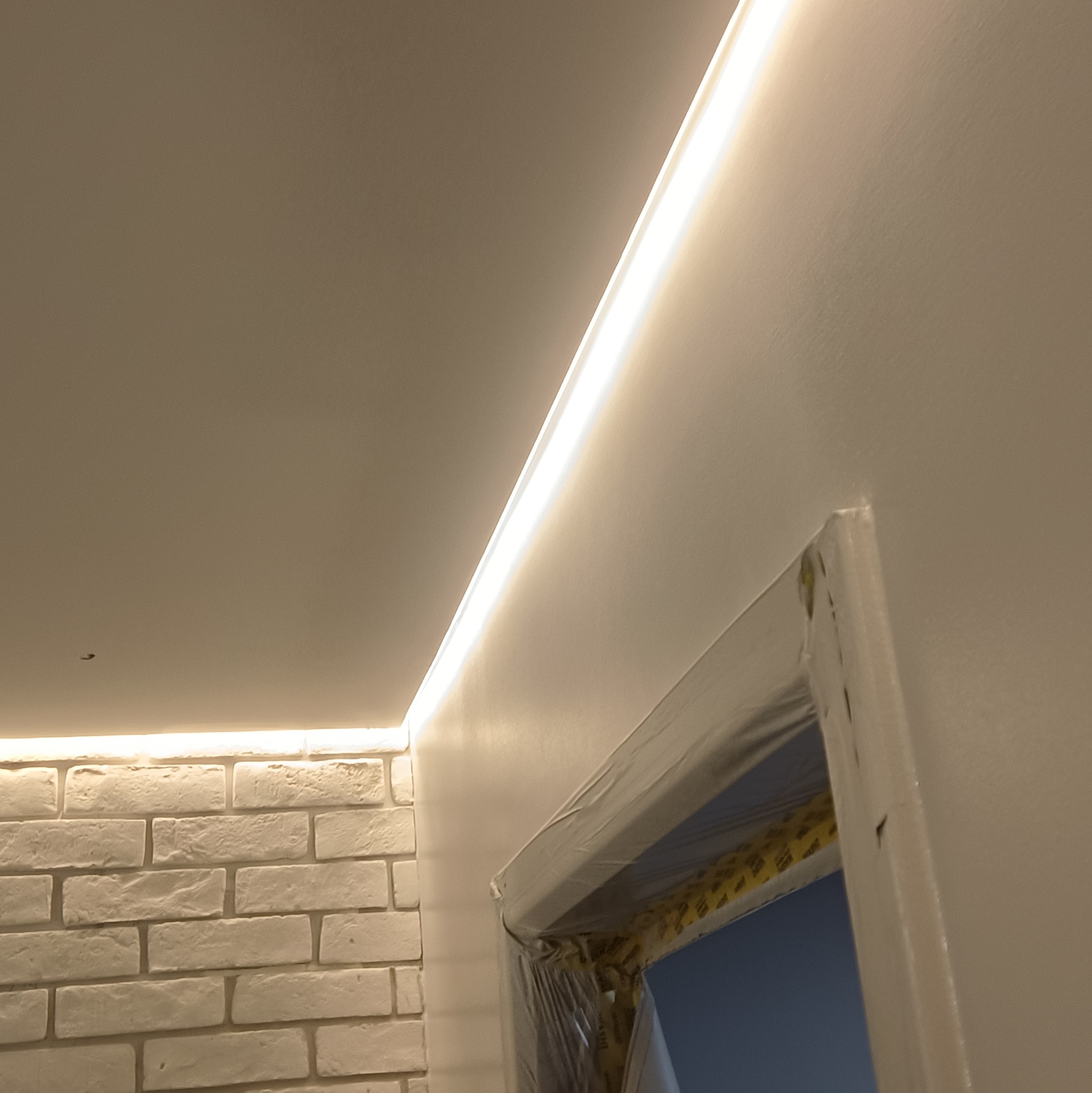 Ściana z cegły i gładzi z oświetleniem LED, zabezpieczone folią drzwi. Detal wykończenia wnętrza z akcentem na nowoczesne oświetlenie i kontrast materiałów.