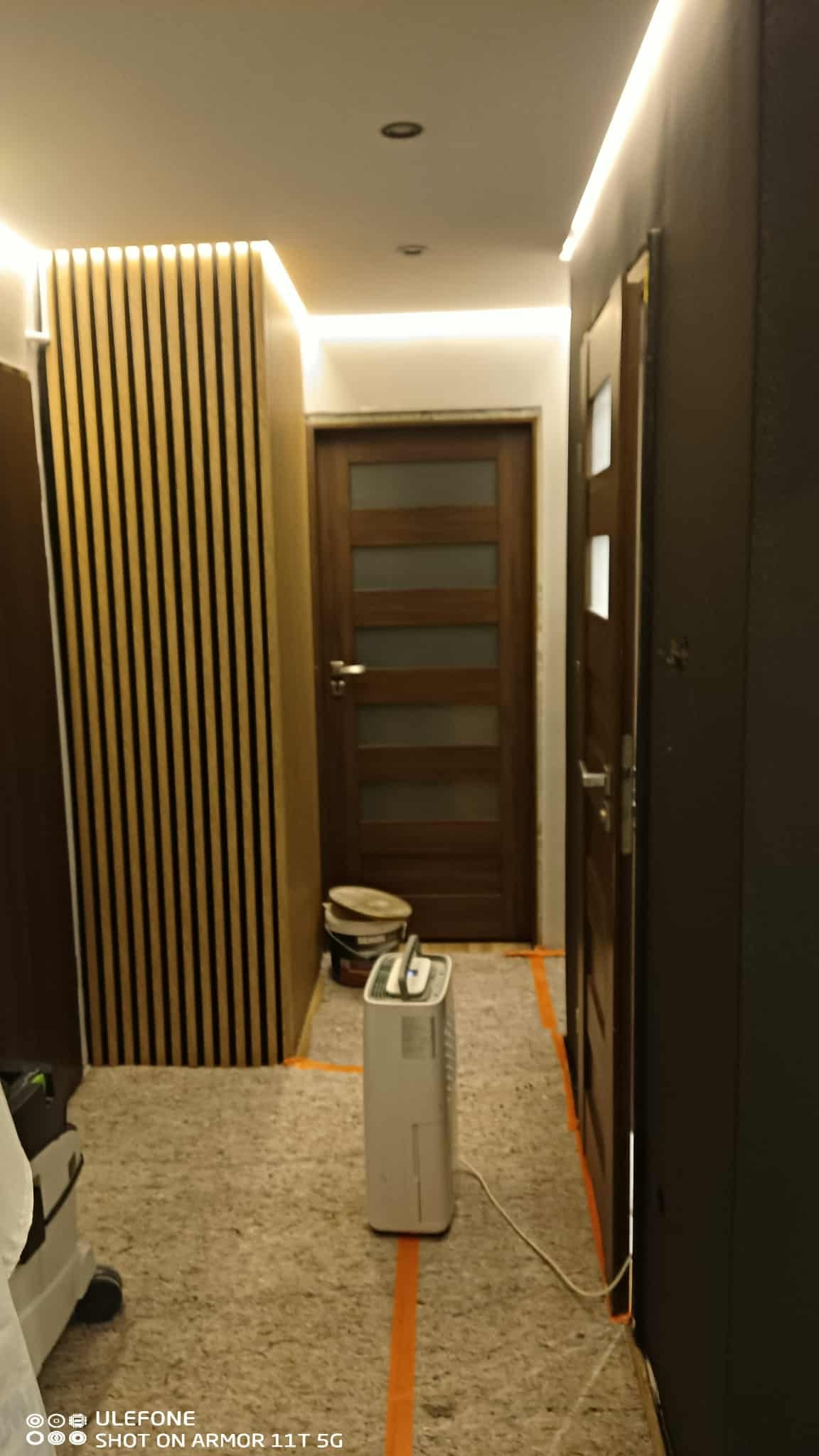 Korytarz po remoncie w Warszawie: drewniane lamele na ścianie, podświetlany sufit, nowe drzwi. Widoczne zabezpieczenia podłogi i sprzęt budowlany.