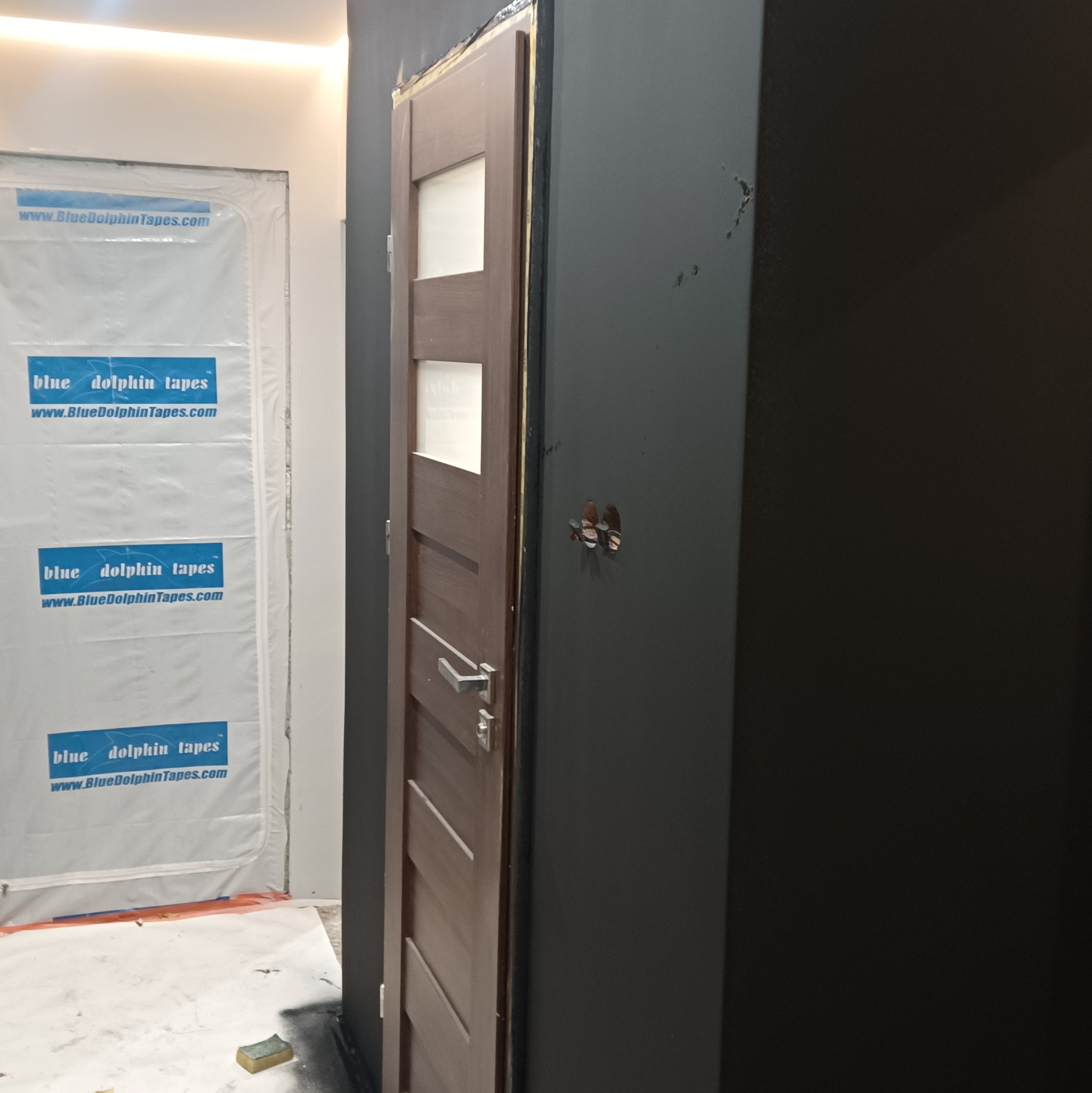 Ciemnoszara ściana po malowaniu z widocznymi śladami po gniazdkach, brązowe drzwi z szybami i folia ochronna na ścianie. Etap prac wykończeniowych.