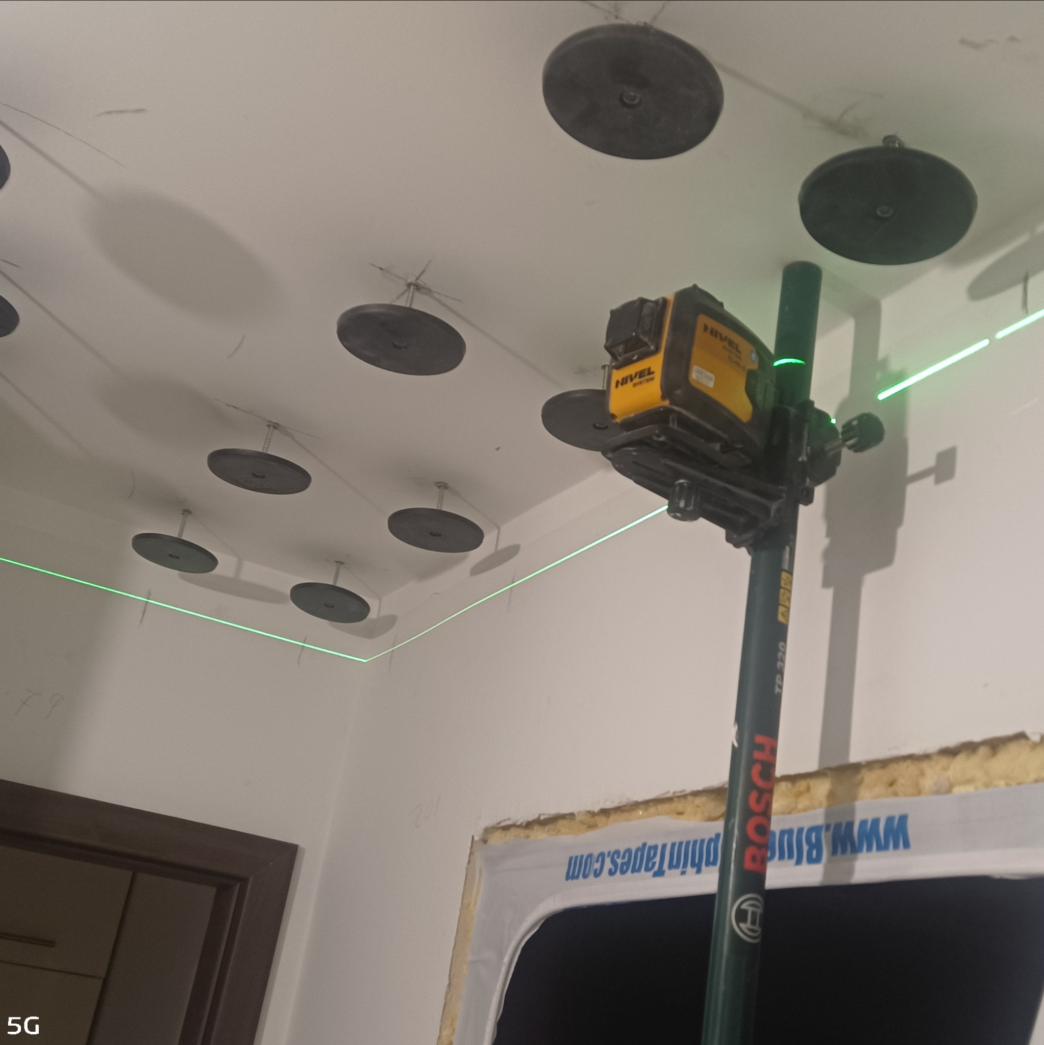 Montaż sufitu podwieszanego: laser krzyżowy wyznacza poziom, widoczne uchwyty montażowe na suficie. Etap prac wykończeniowych, widok z dołu.