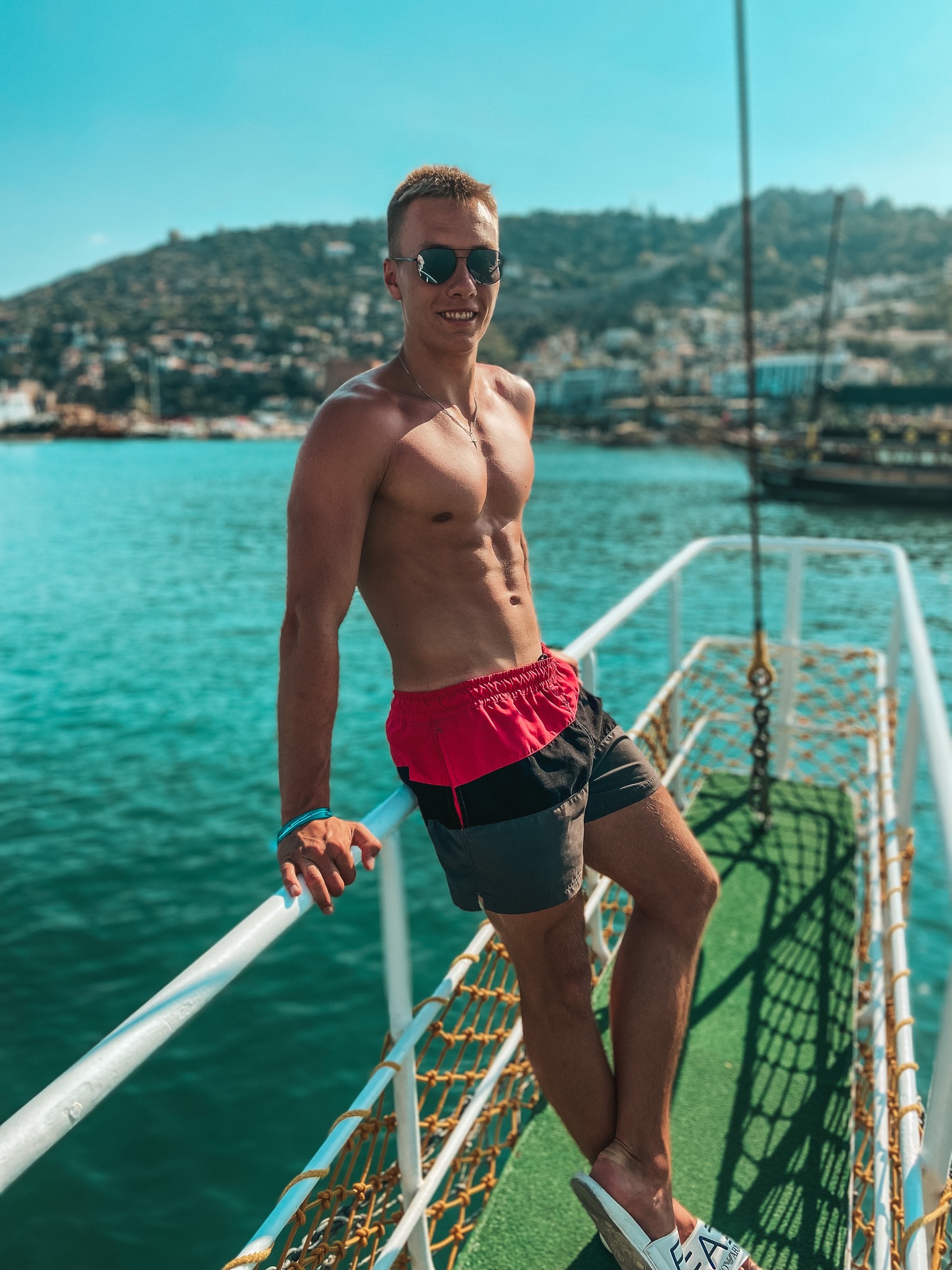 Mężczyzna w okularach przeciwsłonecznych oparty o barierkę łodzi na tle morza i górzystego wybrzeża.