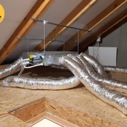 Centrala wentylacyjna z podłączonymi aluminiowymi przewodami w przestrzeni poddasza z drewnianą konstrukcją dachu i płytami OSB, widoczne przewody elektryczne.