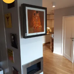 Klimatyzator LG Art Cool Gallery w czarnej ramie, na ścianie w salonie z reprodukcją obrazu 'Pocałunek' Gustava Klimta, obok płyty winylowe i biokominek.