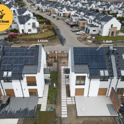 Widok z lotu ptaka na szereg identycznych, nowoczesnych domów z panelami fotowoltaicznymi na dachach; na jednym z domów widoczny napis 5,5kW, na drugim 5,4kW; w lewym górnym rogu logo firmy...