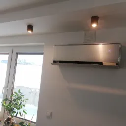 Nowoczesny klimatyzator ścienny z wyświetlaczem temperatury (29 stopni Celsjusza) zamontowany w jasnym pomieszczeniu z trzema okrągłymi lampami sufitowymi i roślinami doniczkowymi na parapecie...