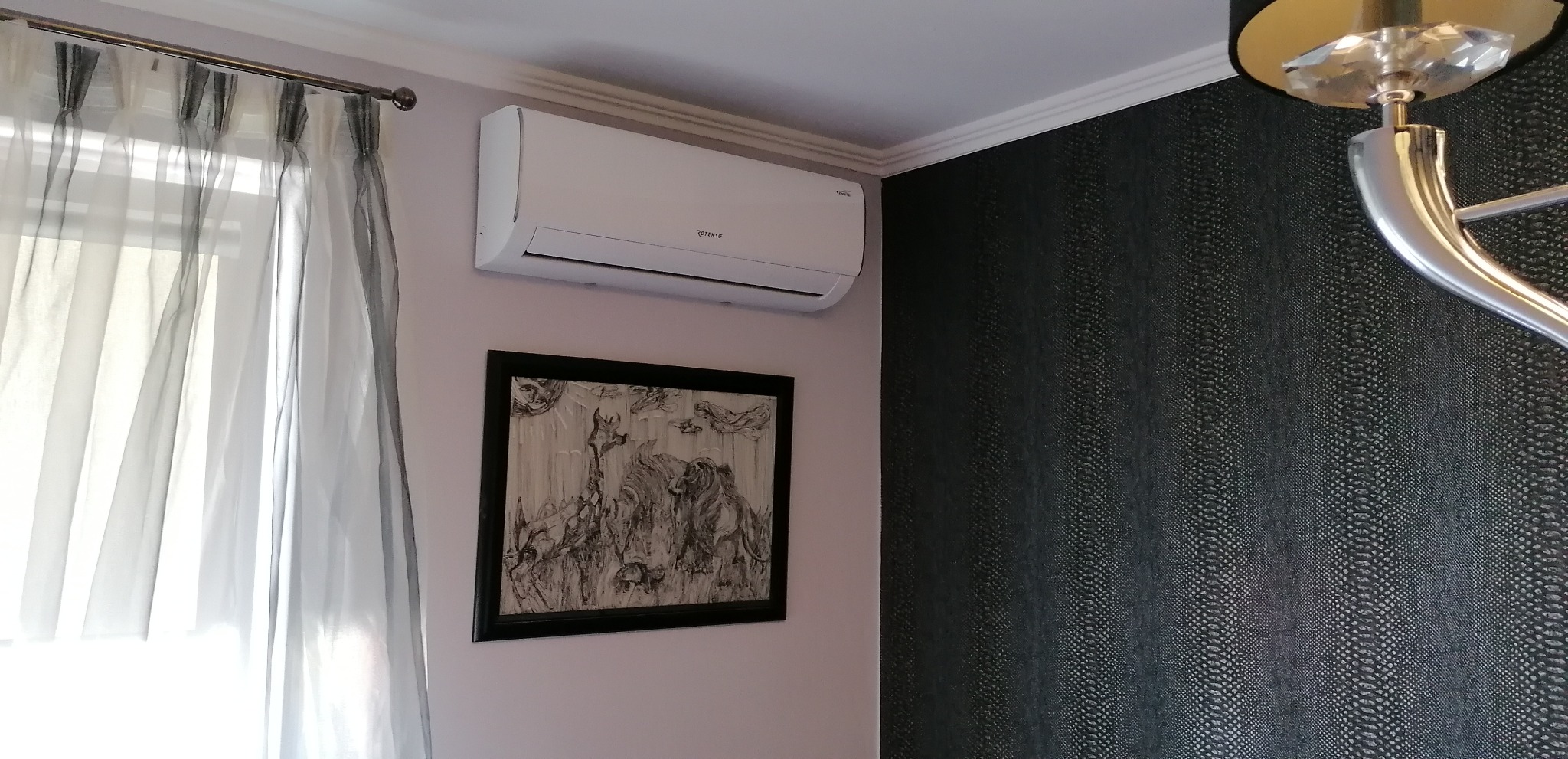 Biały klimatyzator Rotenso zamontowany na ścianie w pokoju, pod nim wisi czarno-biały obraz w ramie, obok okno z firanką i ściana z tapetą imitującą skórę węża.