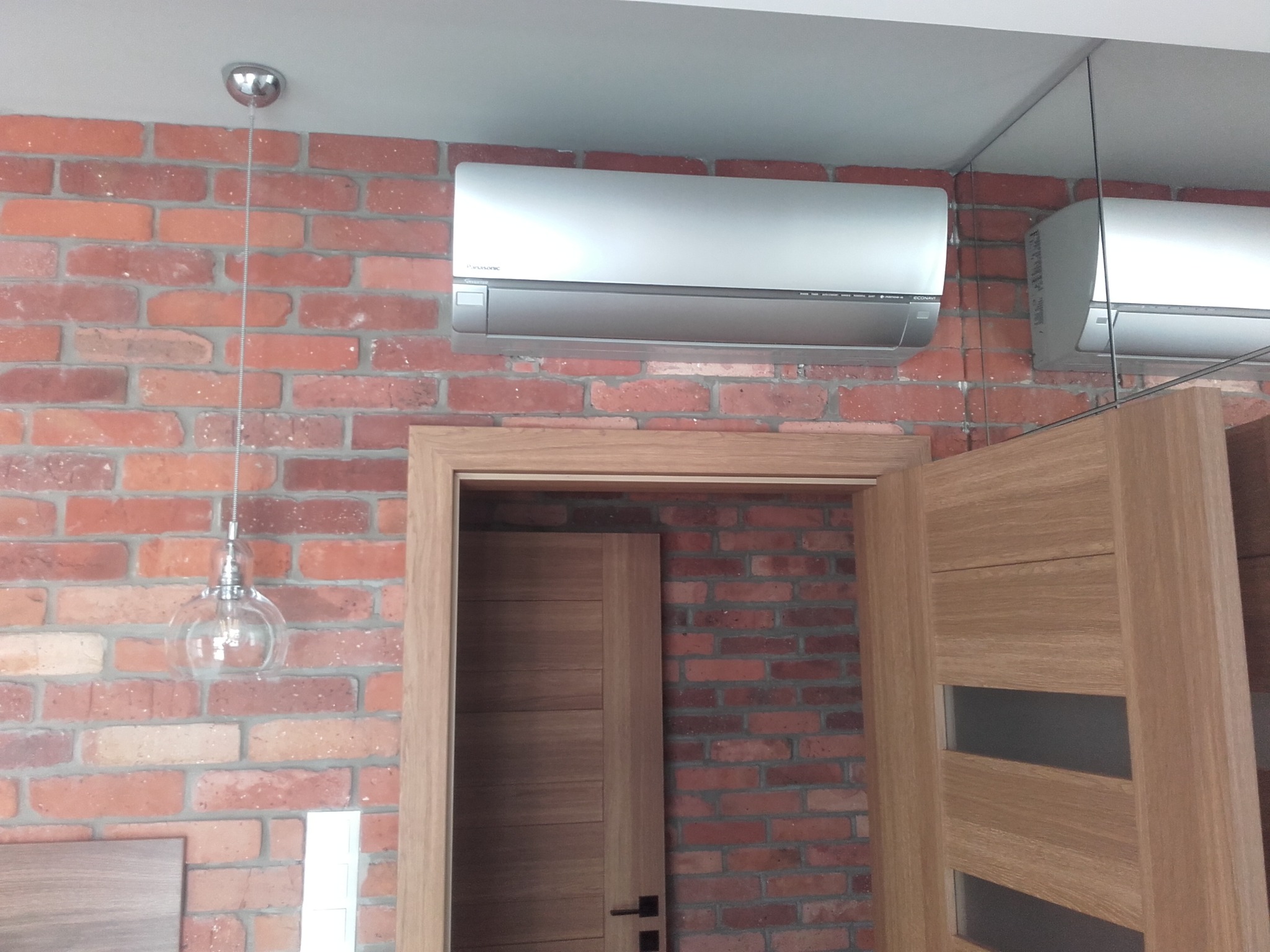 Srebrny klimatyzator zamontowany na ceglanej ścianie w loftowym wnętrzu, widoczna szklana lampa wisząca i fragment drzwi z przeszkleniami.