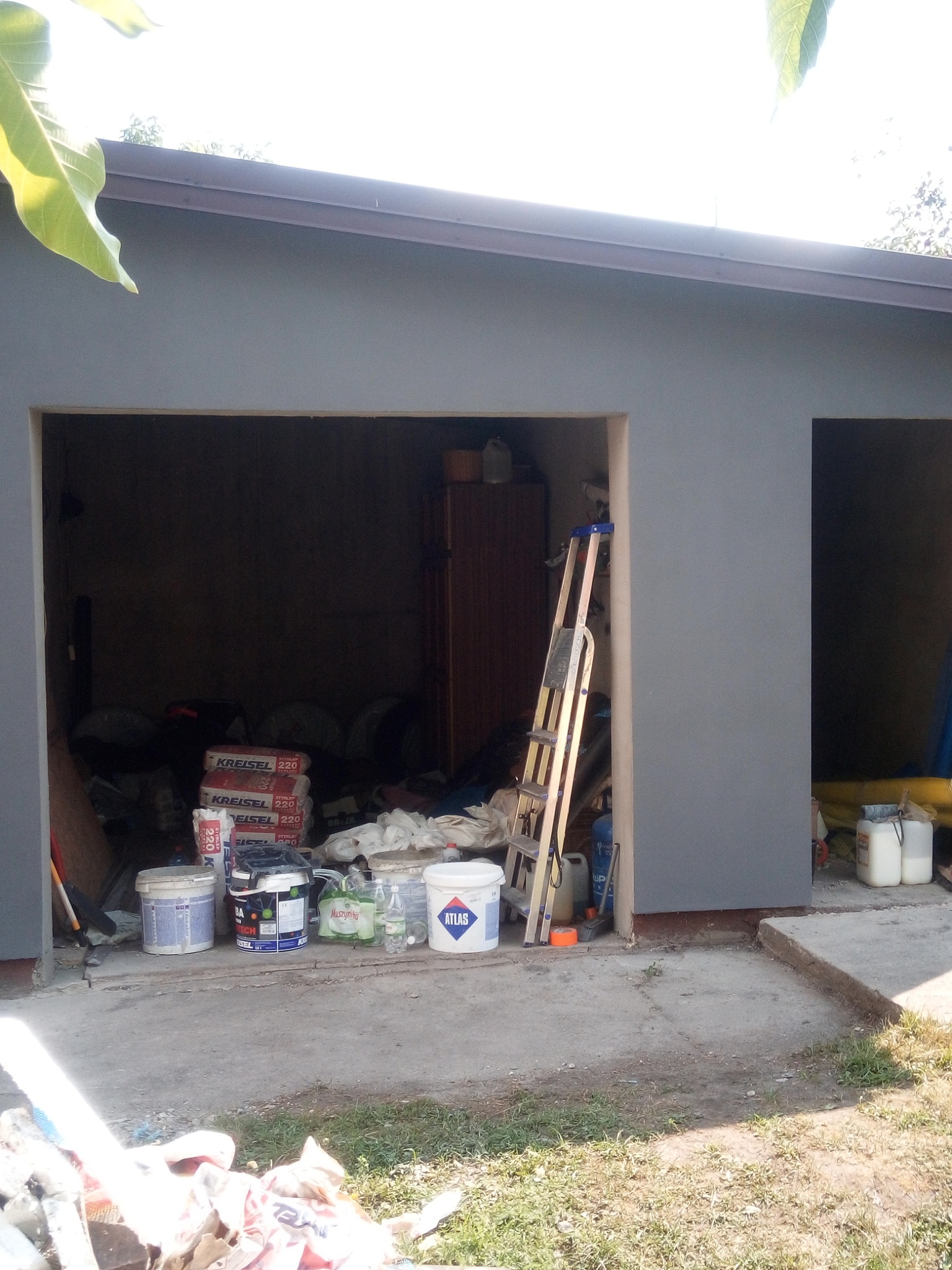 Świeżo pomalowany na szaro budynek gospodarczy z otwartymi wrotami garażowymi, wewnątrz widoczne materiały budowlane, drabina i puszki z farbą różnych producentów, na zewnątrz resztki materiałów...