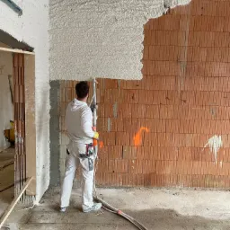 Pracownik w białym kombinezonie aplikuje białą masę na ścianę z cegieł, używając węża i pistoletu natryskowego, widoczne instalacje pod sufitem.