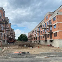 Budowa bloków mieszkalnych w Skoczowie, widoczne surowe mury z czerwonej cegły, okna wstawione, teren budowy w trakcie prac ziemnych, niebieski kontener na gruz.