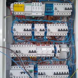 Otwarta skrzynka rozdzielcza z bezpiecznikami i przewodami w różnych kolorach, żółta karteczka z napisem 'NIE ZAŁĄCZAĆ' przy górnym rzędzie aparatury, widoczne elementy marki Legrand i Eaton.