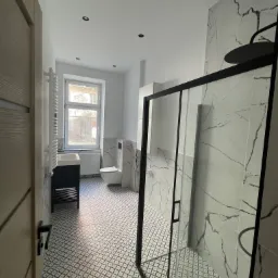 Nowoczesna łazienka z kabiną prysznicową typu walk-in z czarną ramą, marmurowe płytki na ścianach, wzorzysta podłoga, umywalka z szafką i grzejnik drabinkowy.