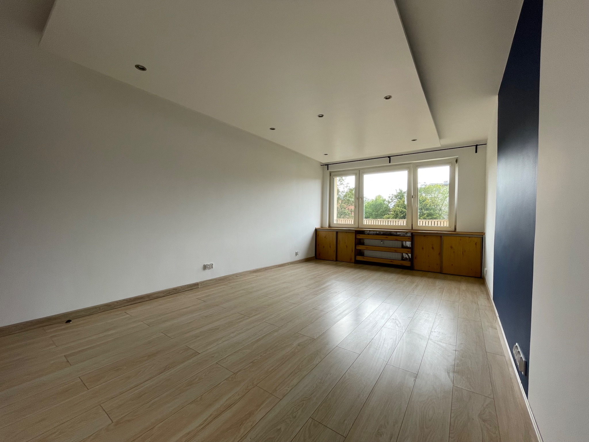 Jasne, przestronne wnętrze po odświeżeniu: parkiet w odcieniach beżu, białe ściany z akcentem granatowej kolumny, zabudowa okienna z drewnianymi szafkami, minimalistyczne oświetlenie sufitowe.