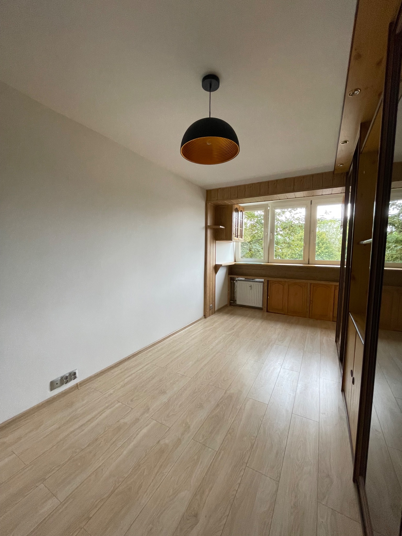 Pusty pokój z jasnymi panelami podłogowymi, białą ścianą z gniazdkami, oknem z drewnianą zabudową i szafą z lustrzanymi drzwiami, oświetlony czarną lampą z miedzianym wnętrzem.