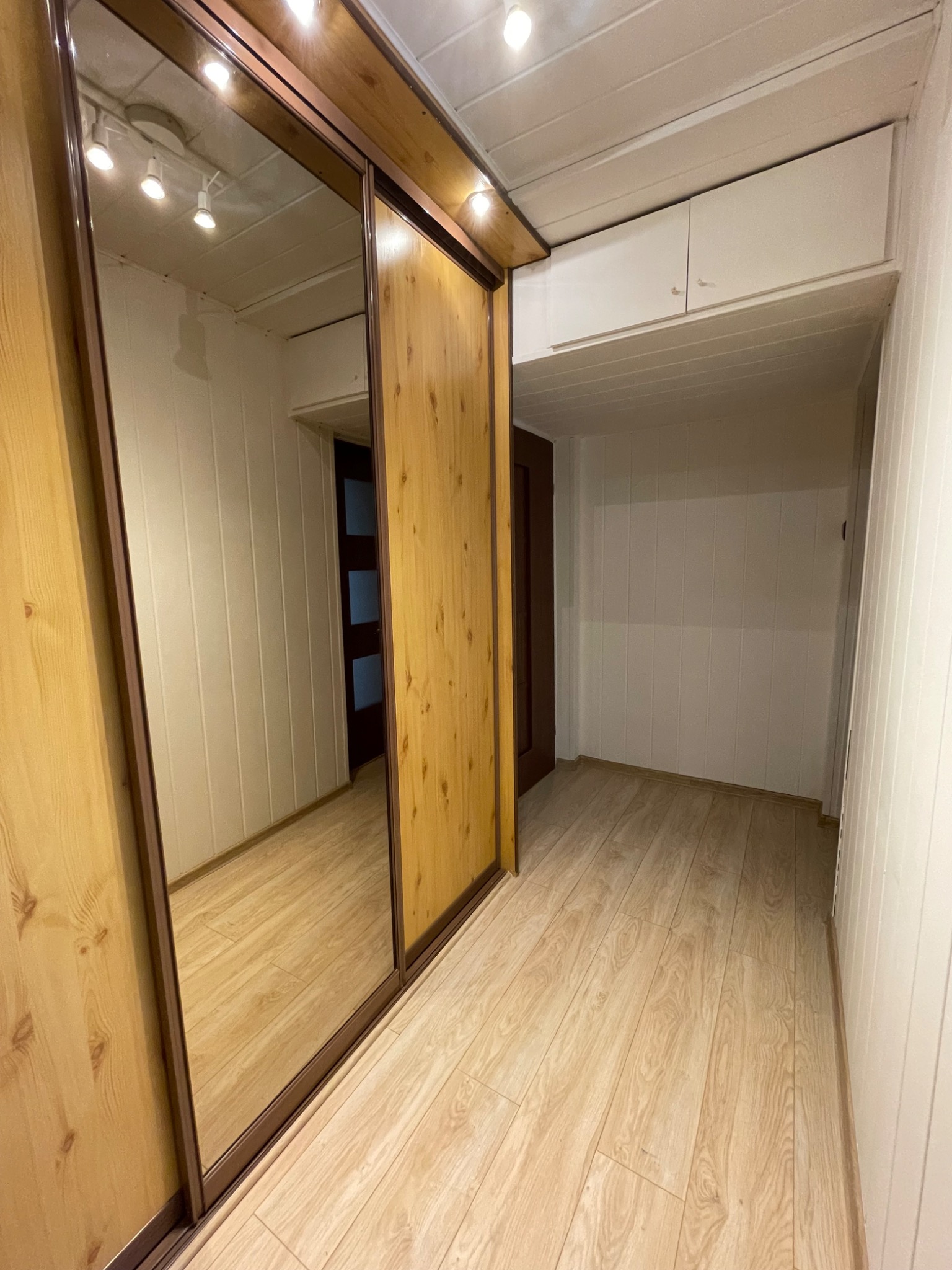 Wnętrze po odświeżeniu, z widoczną szafą przesuwną z lustrem, drewnianą podłogą i białymi panelami ściennymi. W głębi widać otwarte pomieszczenie z dodatkowymi szafkami pod sufitem.