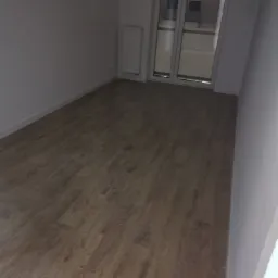 Pusty pokój po remoncie z nowymi panelami podłogowymi o wzorze drewna i świeżo pomalowanymi ścianami na biało, widoczne okno balkonowe.