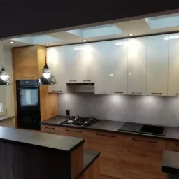 Nowoczesna kuchnia z drewnianymi szafkami dolnymi i białymi górnymi, czarnym blatem, oświetleniem podszafkowym i lampami wiszącymi w stylu industrialnym, widok na wyspę kuchenną.