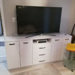 Biała szafka RTV z szufladami i szafkami, na której stoi telewizor; w tle widoczny fragment gitary.