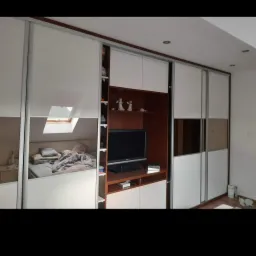 Biała szafa wnękowa z przesuwnymi drzwiami i lustrzanymi panelami, wkomponowana w zabudowę ściany z półkami i miejscem na telewizor.